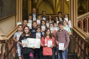 Eine Schulklasse mit etwa 13 Schülerinnen und Schülern präsentiert im Treppenhaus des Erfurter Rathauses gemeinsam mit Oberbürgermeister und den Wanderpokal und die aktuellen Hausaufgabenhefte.