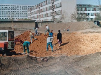 Mehrere Mitarbeiter schaufeln Erde umher um einen Teich zu bauen. Im Hintergrund ein Gebäudekomplex.