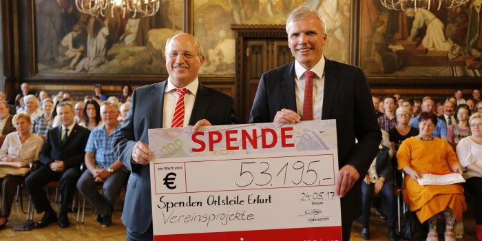 Zwei Herren im Anzug halten einen überdiemensionierten Spendenscheck in Höhe von 53195 Euro in die Kamera, im Hintergrund sieht man einen gut gefüllten Saal. 