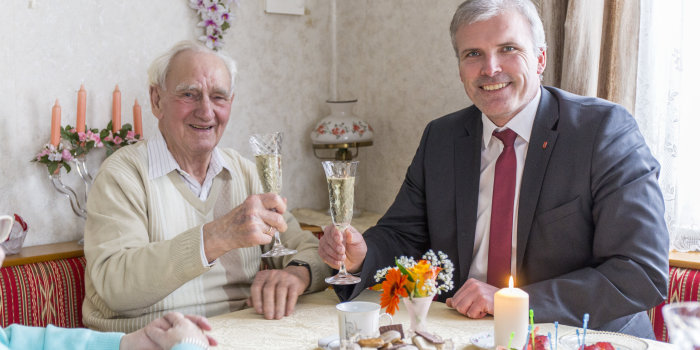 Der Oberbürgermeister und Rudi Keil stoßen mit Sektgläsern im seinem Wohnzimmer am Kaffeetisch auf den Geburtstag Rudi Keils an.