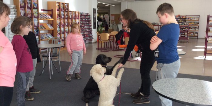 In einem Bibliotheksraum stehen Kinder, vor ihnen zwei Hundem die eine Frau Pfötchen geben.