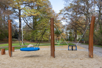 ein Spielplatz mit einer Nestschaukel und einer Schaukel, im Hintergrund ein Karussell für Kleinkinder