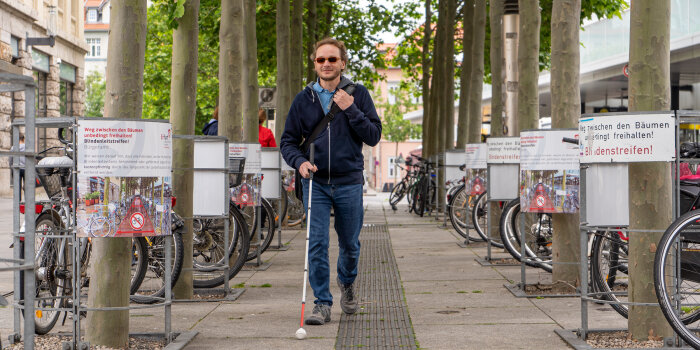 eine sehbehinderte Person mit Blindenlangstock orientiert sich am Blindenleitsystem im Boden