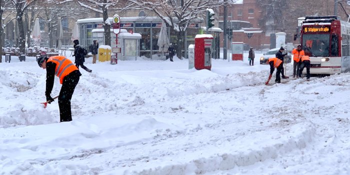 Ein Platz voll mit Schnee, eine Straßenbahn, die in den Gleisen feststeckt, mehrere Personen, die Schnee schippen.