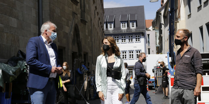 Zwei Männer und eine Frau stehen auf einer Straße, im Hintergrund sind Dreharbeiten zu sehen.