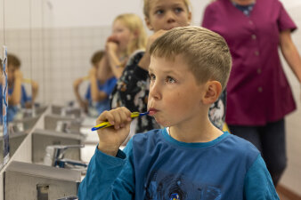 Ein Junge putzt sich die Zähne. Im Hintergrund stehe weitere Kinder und eine Frau.