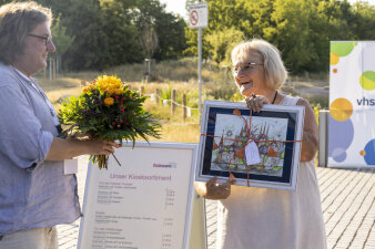 eine Frau hält ein gerahmtes Bild in der Hand, am linken Bildrand hält ein Mann Blumen in der Hand