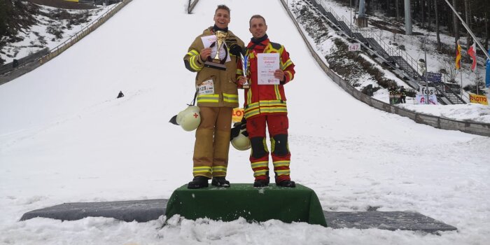 Zwei Männer in Feuerwehrkleidung stehen im Auslauf einer Skisprungschanze.