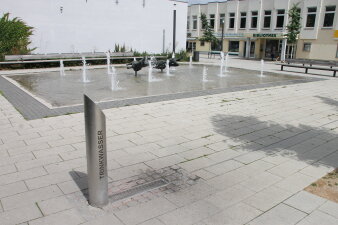 Ein stählerner Trinkbrunnen als Säule auf einer gepflasterten neben einem Springbrunnen.