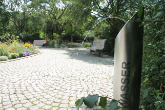 Ein stählerner Trinkbrunnen als Säule auf einer gepflasterten Fläche in einem Park. 