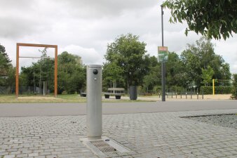Ein stählerner Trinkbrunnen als Säule auf einer gepflasterten Fläche in einem Park. 