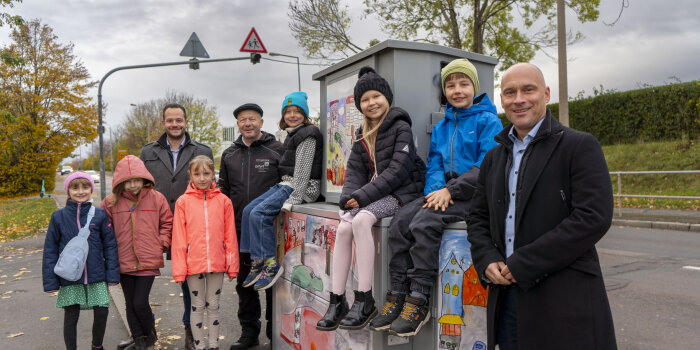 Eine Gruppe von Kindern und drei erwachsene Männer stehen lächelnd vor einem Gerät zur Geschwindigkeitsmessung.