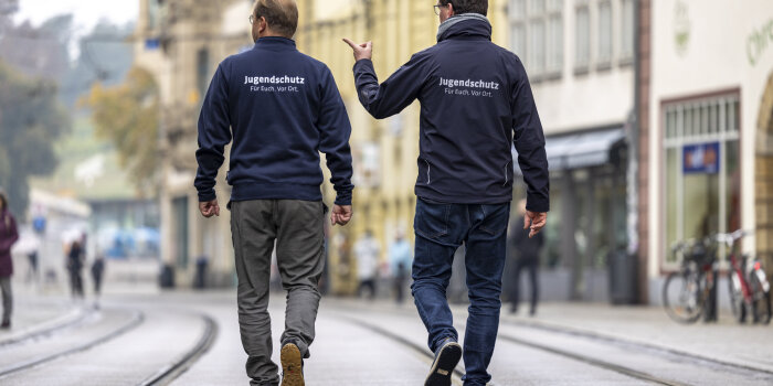 Rückansicht zweier Männer, die in Dienstkleidung durch eine Straße laufen und gestikulieren.