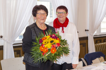 Zwei ältere Frauen stehen beisammen. Beide tragen eine Brille und eine hält einen bunten Blumenstrauß in der Hand.
