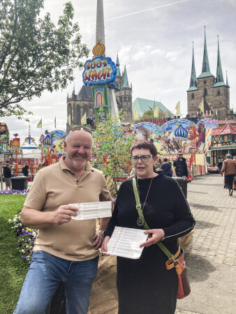zwei Personen halten bedrucktes Papier in der Hand, im Hintergrund ein Volksfest in Erfurt