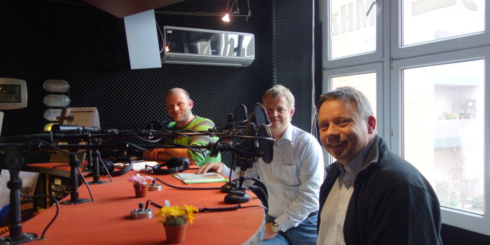 Drei Herren sitzen am Mikrofon in einem Hörfunkstudio.
