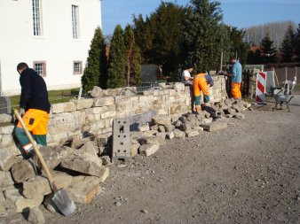 Vier Fachkräfte haben eine mannshohe Mauer fast fertig gebaut. Es liegen noch Steine und Schaufeln herum und sind teilweise in Benutzung.
