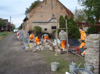 Sechs Fachkräfte arbeiten in ihren meist orangenen Arbeitsklamotten an dem Aufbau einer Mauer. Das geschieht im Umfeld einer kleinen dörflichen Wohngegend, Absperrungen sind vorhanden.