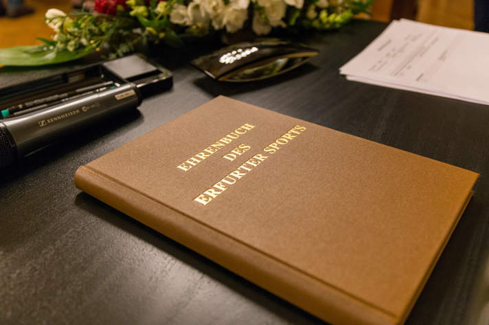 Ein in braunen Stoff gebundenes Buch mit dem Titel „Ehrenbuch des Erfurter Sports“ liegt auf einem dunklen Tisch, ein Mikrofon, Blumen sowie Blätter liegen daneben.