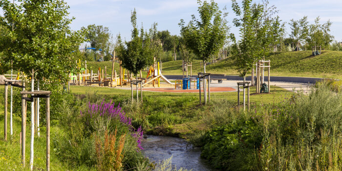 Blick über einen Bach in eine Parklandschaft mit einem Spielplatz