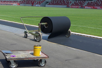 In einem Stadion wird auf einem Asphaltboden ein schwarzer Kunststoffbelag ausgerollt.