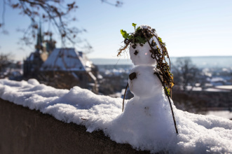 Im Vordergrund zu sehen ist ein kleiner Schneemann auf einem Mauersims des Petersberges, im Hintergrund ein Blick über die Stadt und den Dom.