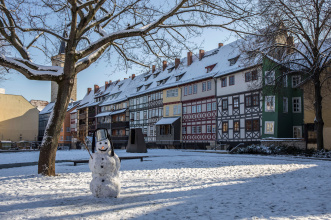 Im Vordergrund ist ein Schneemann mit einem Metalleimer auf dem Kopf zu sehen, stehend auf einer verschneiten Wiese mit kahlen Bäumen. Im Hintergrund die Häuser der Krämerbrücke von außen mit ihren zugeschneiten Dächern.