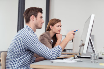 Ein mittdreißiger Mitarbeiter zeigt seiner gleichaltrigen Kollegin an einem Computerbildschirm Sachen auf. Ein Ordner mit Unterlagen liegt vor ihnen.