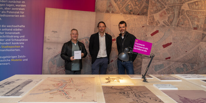 drei Männer stehen in einer Ausstellung, vor ihnen ein Tisch mit verschiedenen Plänen und Abbildungen