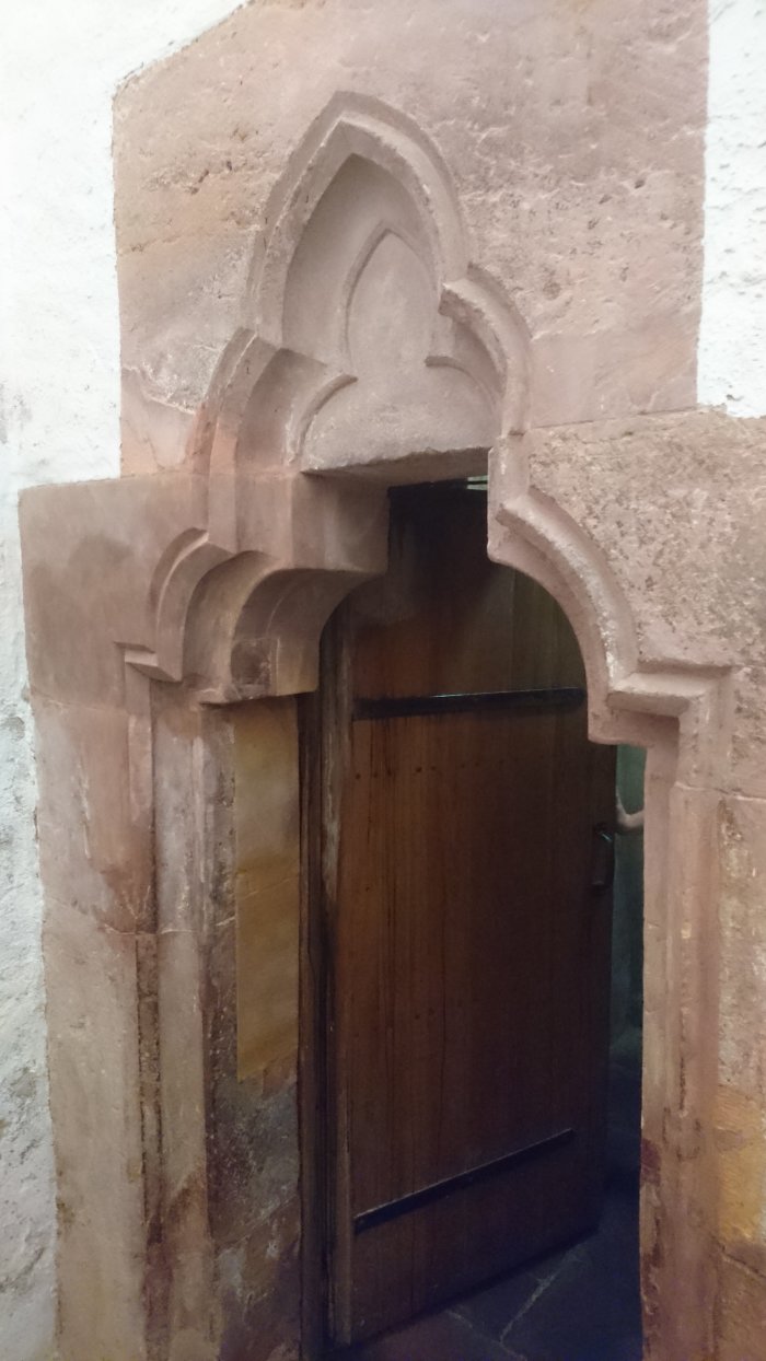 Zu sehen ist ein schmales gotisches Eingangsportal mit spitzbogigem steinernen Dekor. Dir geöffnete Holztür geht nach innen auf, der Betrachter steht vor dem Eingang, in den Raum hinein sieht man jedoch nicht.