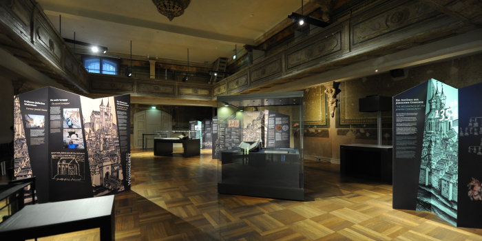 Blick vom Treppenhaus aus in einen Raum mit Holzboden, in dem Ausstellungstafeln und Vitrinen zu sehen sind