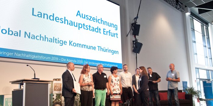 Foto: Global Nachhaltige Kommune © Sylwia Mierzynska Menschen auf einer Bühne, dahinter eine Projektionswand.