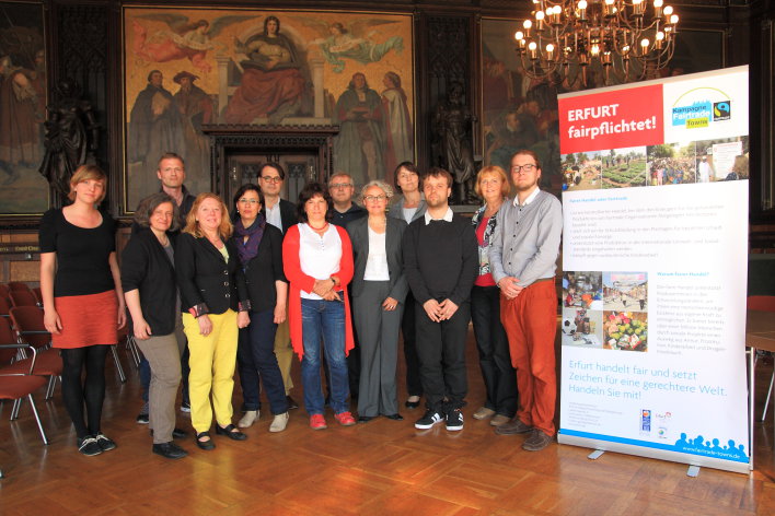 Foto: Steuerungsgruppe Fairtrade Town Erfurt © Stadtverwaltung Erfurt / J. Ludwig Dreizehn Personen im Halbkreis aufgestellt, rechts ein Roll-on-Plakat mit der Aufschrift "Erfurt fairpflichtet! – Kampagne Fairtrade Towns"