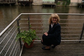 Eine blonde Frau hockt auf einem Balkon vor einem mit frischem Grün bepflanzten Blumentopf