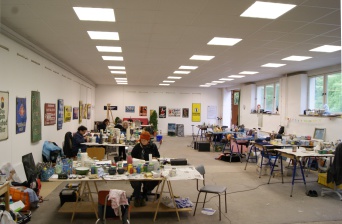 Ein großer Arbeitssaal. An den Tischen sitzen einzelne Künstler. An den Wänden Emailleschilder. Insgesamt ein recht buntes Bild.