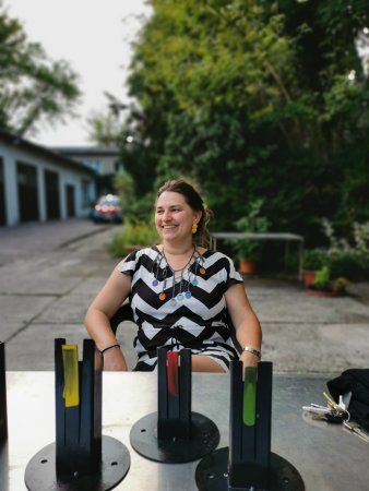 lächelnde junge Frau mit Halsschmuck, im Vordergrund auf einem Tisch Arbeiten aus emailliertem Stahl