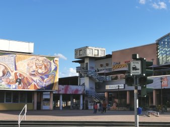 Eingang zu Einkaufspassage, links große Wandmalerei, in der Mitte Betonturm, rechts Geschäfte