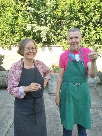 links Frau mit schwarzer Brille und Schürze über rosa-lila-gelb gemusterter Bluse, rechts Mann mit pinkem T-Shirt