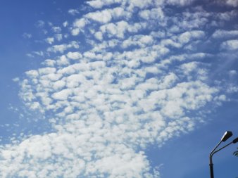 wattebauschartige Wölkchen in Reihen bilden zwei ovale Formen am Himmel, rechts unten Detail einer Straßenlaterne