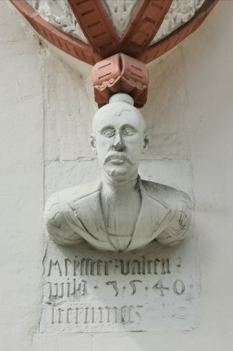 erkertragende weiße Schulterkopffigur, oben ein rotes Ornament, unten Schrift