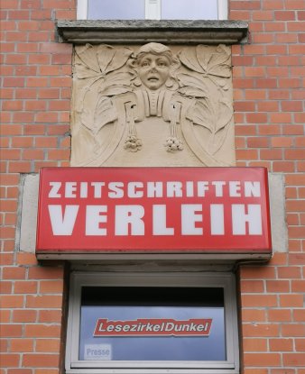 Jugendstil-Steinrelief mit unglücklichem Frauengesicht, darunter ein Firmenschild mit Text "Zeitschriften Verleih"