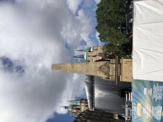 Ein brauner Obelisk vor einer Kirchenkulisse, dahinter blauer Himmel