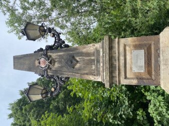 Ein brauner Obelisk mit zwei Lampen vor Bäumen und blauen Himmel