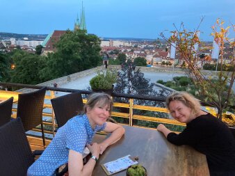 Zwei lächelnde Frauen vor einer Stadtsilhouette mit Bäumen im Hintergrund