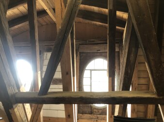 Blick auf verschiedene Holzbalken in einem Dachstuhl, im Hintergrund zwei Fenster durch die Licht fällt