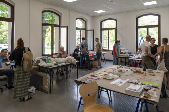 Tag der offenen Tür bei 19. Erfurt Schmucksymposium 2023 Ein heller Raum mit vielen Fenstern, darin mehrere Tische mit Objekten und Personen die sich unterhalten