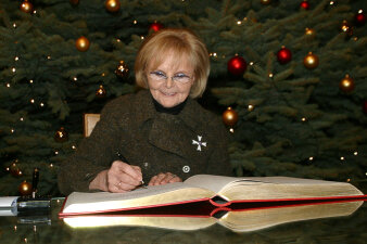 Eine Frau sitzt an einem Tisch und schreibt sich in ein Ehrenbuch ein, im Hintergrund steht ein Weihnachtsbaum.