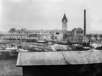Schwarz-Weiß-Foto eine Industrieanlage. Auffällig sind ein gemauerter Turm und ein Schornstein. 