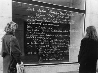 Schwarz-Weiß-Foto eines Schaufensters mit Beschriftung. Davor stehen zwei Frauen und lesen. 