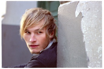 Junger Mann mit blonden Haaren, links im Bild, leht an einer Mauer.